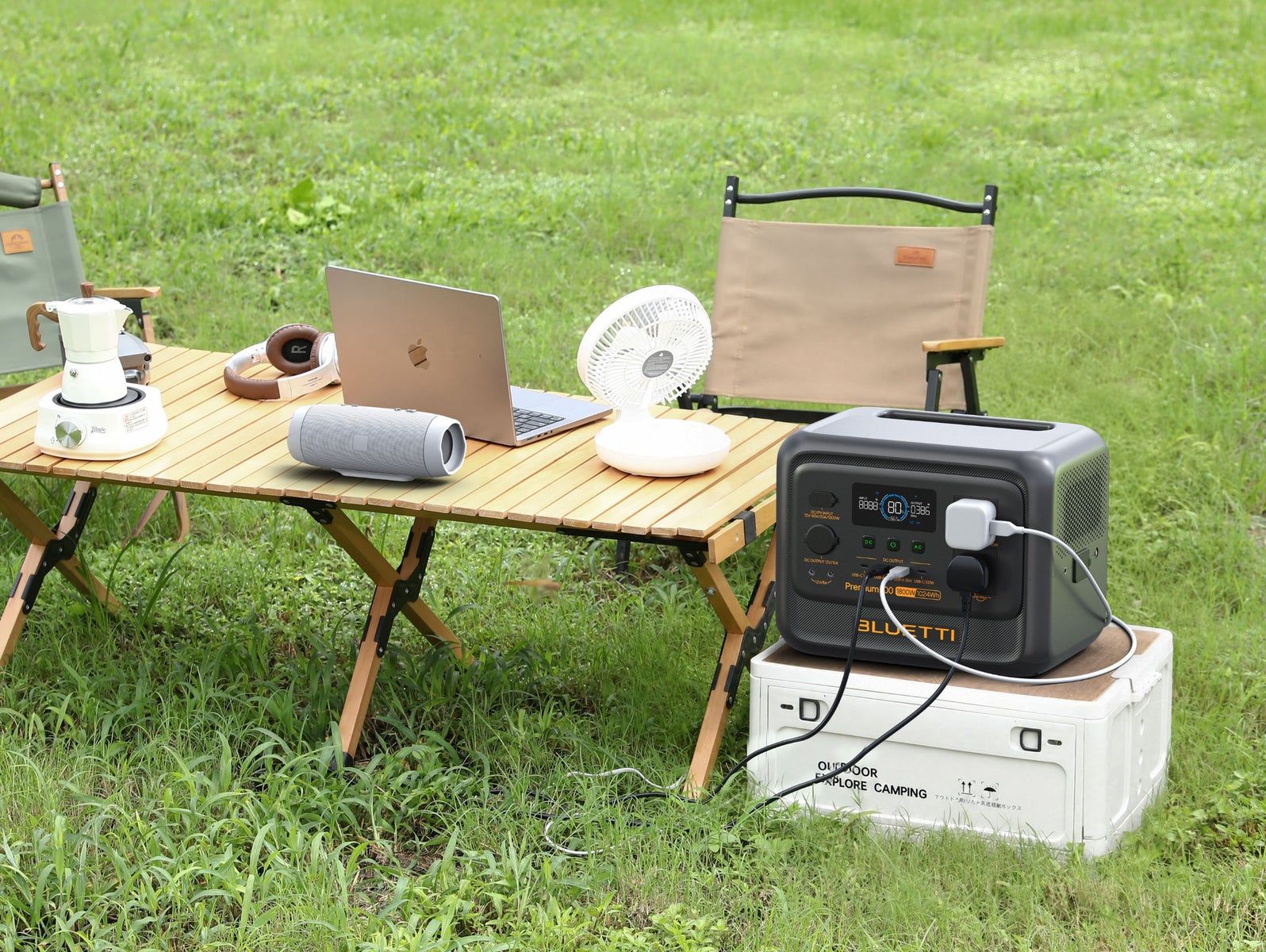 Camp table on grass with moka pot on a hotplate, speaker, laptop, desk fan, headphones, Premium 100 V2 on a storage box running them, a white UK USB charger and a black UK plug in its two AC outlets