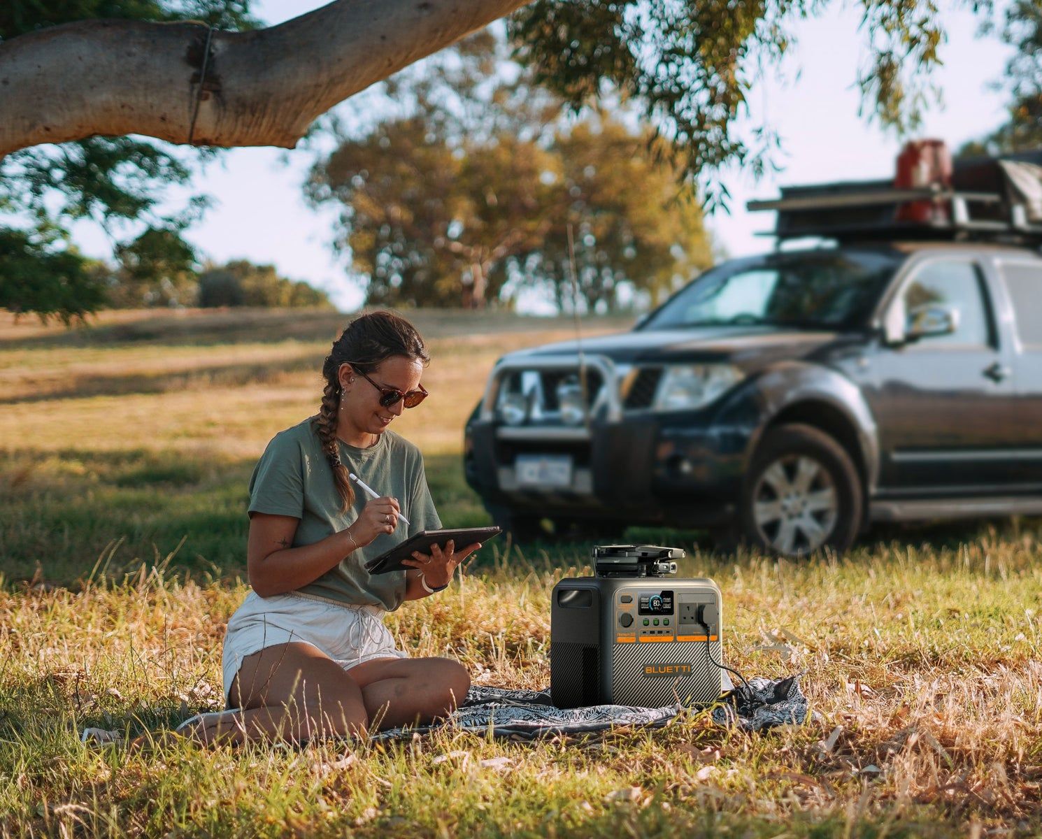 Woman with a tablet sitting on grass beside Premium 200 V2, pickup with roof tent behind